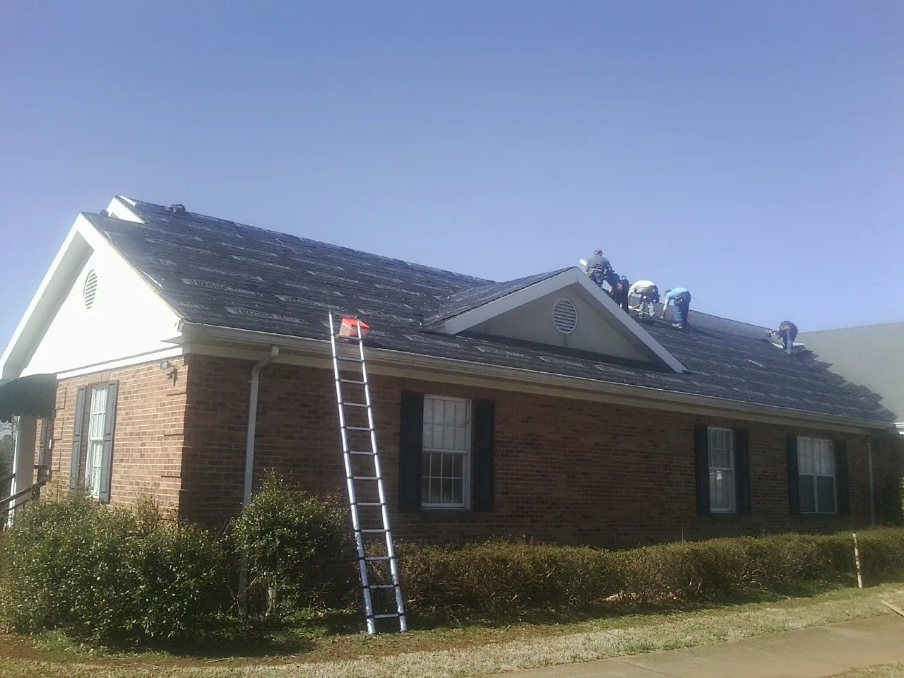 Crew tearing shingles off a brick ranch roof with a ladder set — Emergency Roof Repair work by our crew in Flovilla