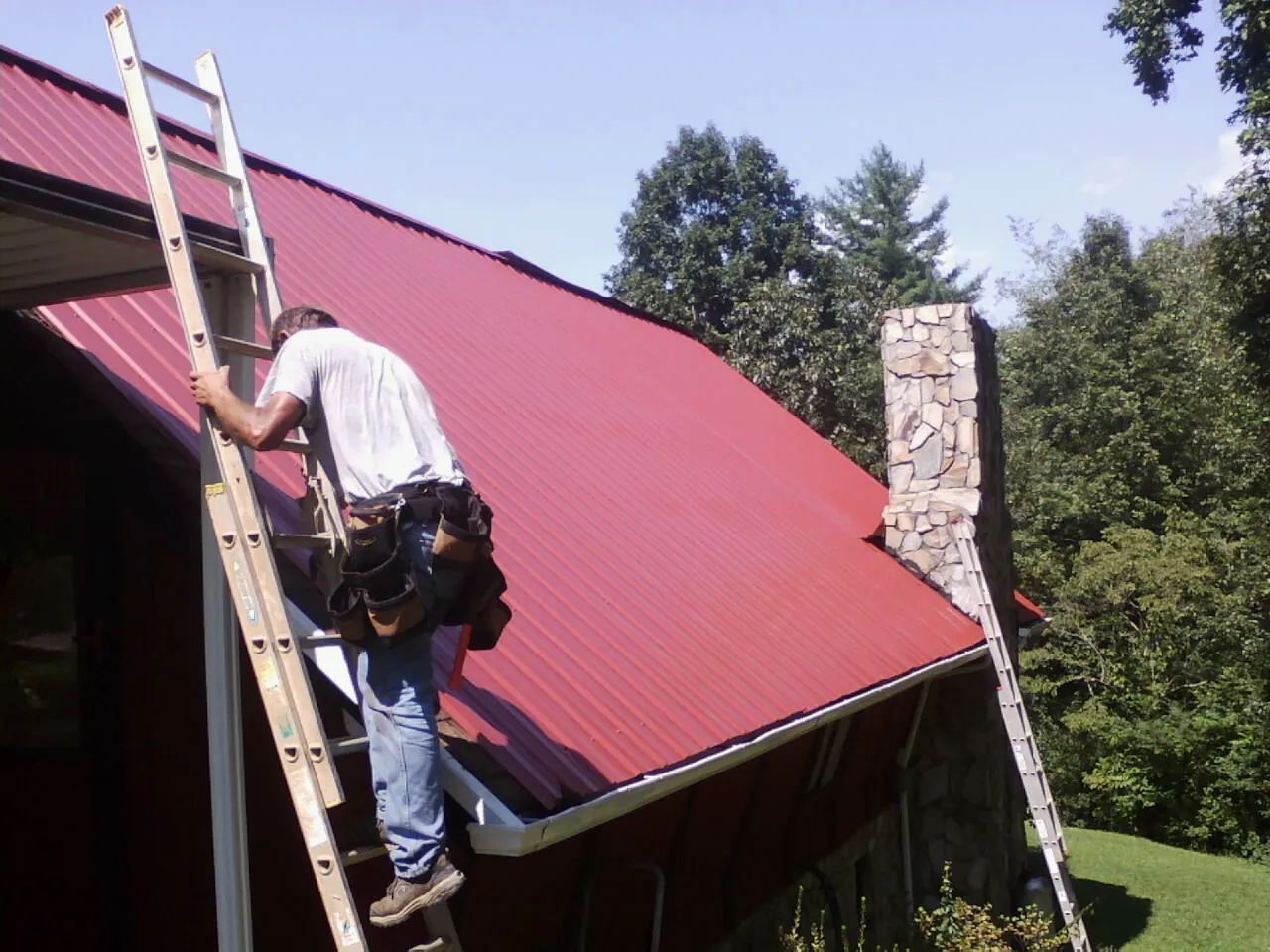 Roofer on a ladder beside a red metal roof and stone chimney — Soffit & Fascia Repair by Roofing Brothers in Flovilla
